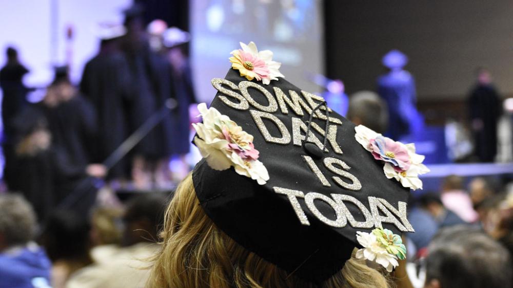 Uno studente è seduto Dominican Universitycerimonia di inizio. La schiena dello studente è rivolta alla telecamera. Il berretto da laurea dello studente è decorato con fiori e la scritta Some Day is Today.
